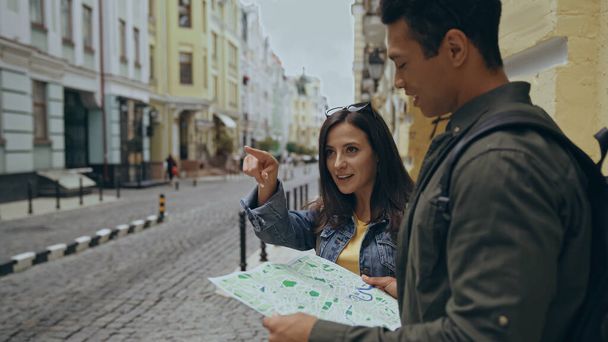 Smiling tourist pointing with finger near bi-racial boyfriend with map on urban street  - Foto, Bild