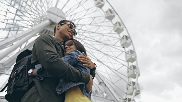 Low angle view of bi-racial tourist hugging girlfriend near ferris wheel  - Valokuva, kuva