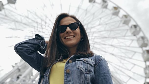 Low angle view of smiling tourist in sunglasses standing near blurred ferris wheel  - Photo, Image