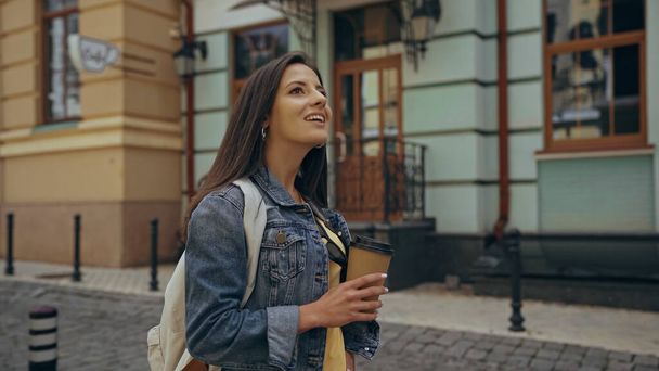 Smiling tourist with backpack holding takeaway drink on urban street  - Zdjęcie, obraz