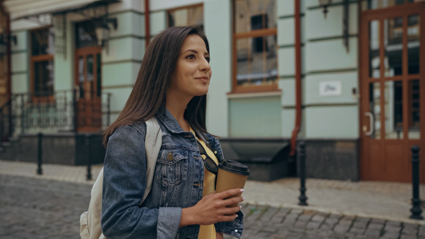 Positive tourist with backpack holding coffee to go on urban european street  - Zdjęcie, obraz