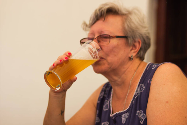 Smiling caucasian senior woman with gray hair holding and drinking glass of light beer in craft czech brewery. Alcoholic drink. Weekend meeting with friends in pub. Oktoberfest concept. - Photo, Image