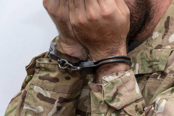 A convicted military man in handcuffs covers his face with his hands on a light background. Concept: war criminal, army court and tribunal, sentencing for a prisoner. - Foto, Bild