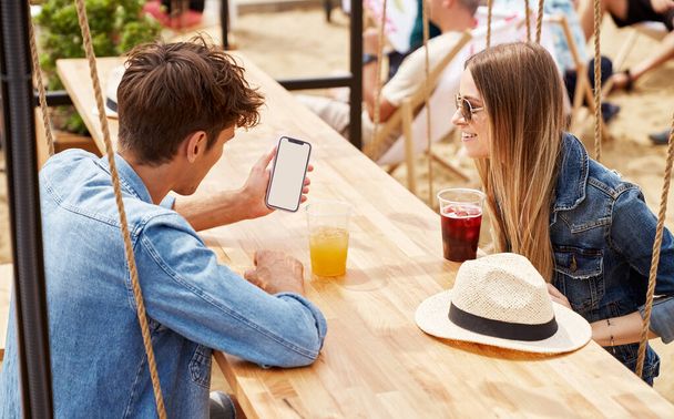 Young couple of freinds sitting drinking ice juice taliking and looking at blank screen of smartphone  - Photo, Image
