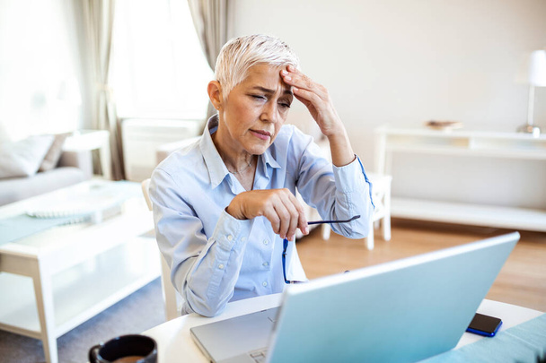 Senior frustrated woman working from home office in front of laptop, having a headache.Mature woman having a painful face expression - Photo, image