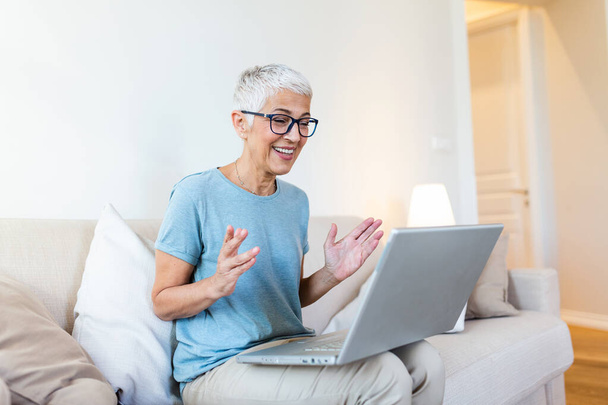 Glückliche reife Frau winkt jemandem zu, während sie zu Hause einen Videoanruf über den Laptop führt. Grauhaarige Seniorin fuchtelt mit der Hand vor dem Laptop, während sie Videotelefonie mit ihren Familienmitgliedern führt. - Foto, Bild
