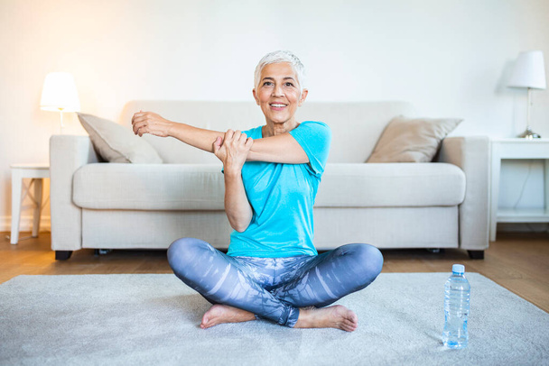 Senior woman doing warmup workout at home. Fitness woman doing stretch exercise stretching her arms - tricep and shoulders stretch . Elderly woman living an active lifestyle. - Photo, Image