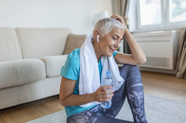 Senior woman holding plastic bottle of water,wiping sweat with a towel, exhausted after the daily training. Elderly woman taking a break while exercising at home. sitting on fitness mat and resting after training at home - 写真・画像