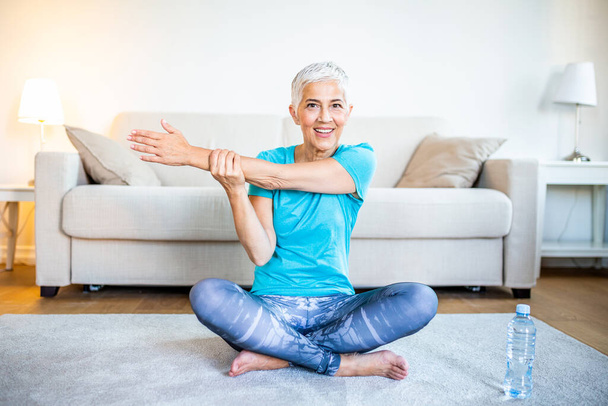 Senior woman doing warmup workout at home. Fitness woman doing stretch exercise stretching her arms - tricep and shoulders stretch . Elderly woman living an active lifestyle. - Foto, afbeelding