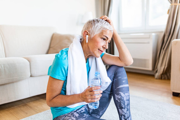 Senior woman holding plastic bottle of water,wiping sweat with a towel, exhausted after the daily training. Elderly woman taking a break while exercising at home. sitting on fitness mat and resting after training at home - Foto, afbeelding