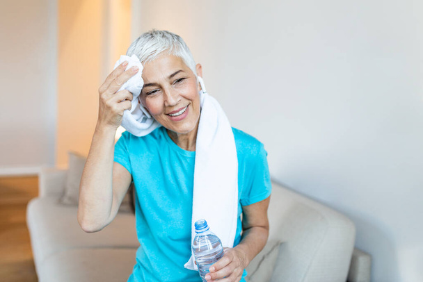 Senior woman holding plastic bottle of water,wiping sweat with a towel, exhausted after the daily training. Elderly woman taking a break while exercising at home. sitting on the sofa,smiling and resting after training at home - Foto, afbeelding