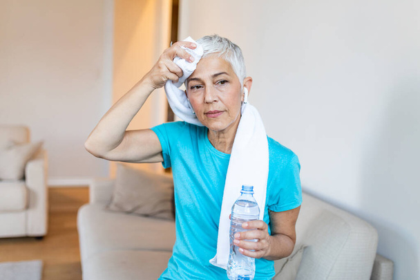 Senior woman holding plastic bottle of water,wiping sweat with a towel, exhausted after the daily training. Elderly woman taking a break while exercising at home. sitting on the sofa and resting after training at home - Fotografie, Obrázek