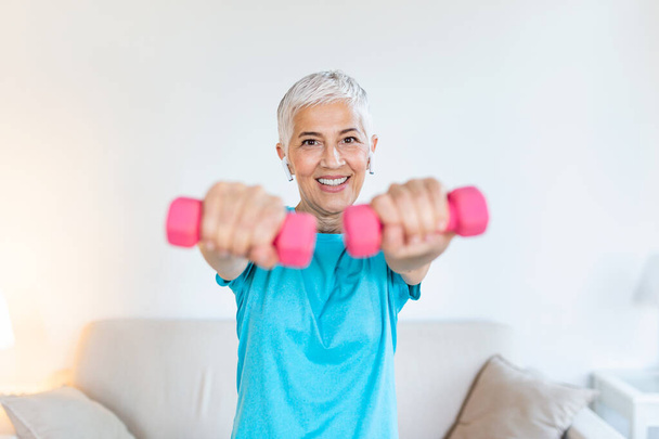 Senior woman exercising with dumbbells at home. Sporty beautiful woman exercising at home to stay fit. Elderly woman exercising at home in a living room. Fitness, workout, healthy living and diet concept. - 写真・画像