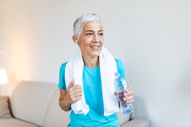 Senior woman with headphones while resting after workout. Having a towel around her neck. Holding a bottle of water. Athletic mature woman resting after a good workout session - Photo, Image