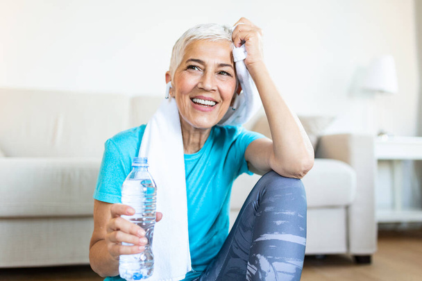Senior woman holding plastic bottle of water,wiping sweat with a towel, exhausted after the daily training. Elderly woman taking a break while exercising at home. sitting on fitness mat and resting after training at home - Fotografie, Obrázek