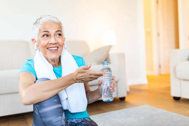 Senior woman with headphones while resting after workout. Holding a bottle of water. Athletic mature woman resting after a good workout session - Foto, Imagen