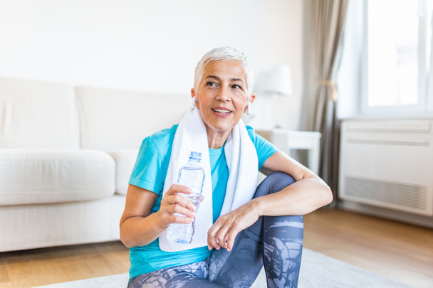 Elderly woman sitting on the mat, exhausted after the daily training.Senior woman taking a break while exercising at home. Athletic mature woman holding bottle of water - Foto, Bild