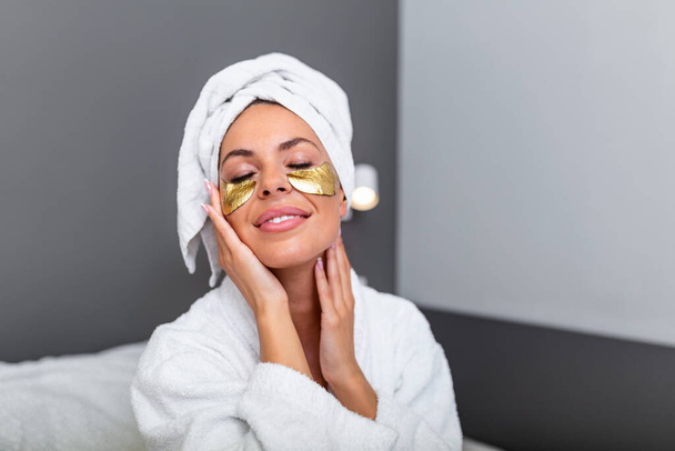 Beautiful young woman with Gold Cosmetics mask, Eye Patches. sitting on bed in white bathrobe in bedroom at home. Smiling young woman lying on bed in bathrobe relaxing on the bed after bath - Valokuva, kuva