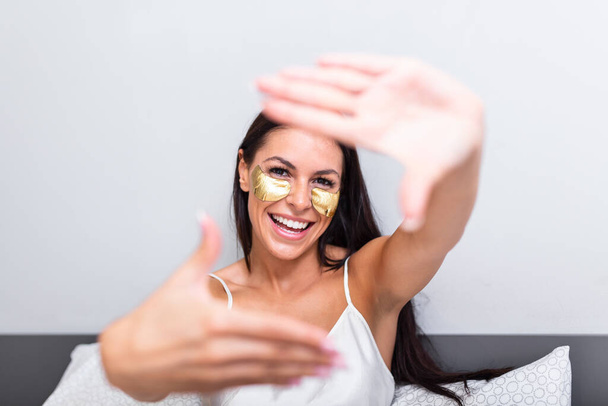 Image of woman with Gold Cosmetics mask, Eye Patches. making photo frame sign with fingers while lying in bed. Portrait of smiling lady posing in bright home apartments. Concept happiness and love. - Valokuva, kuva