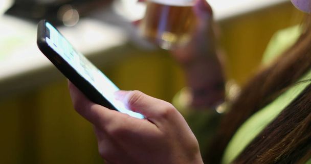Closeup of young woman hand using cellphone touchscreen while drinking alcholic beverage at bar nightlife - Photo, image