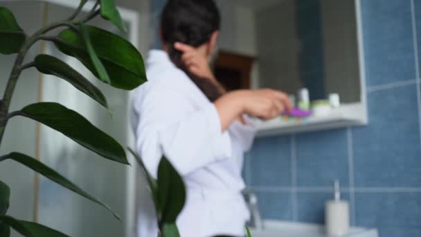 View through houseplant: Curly brunette, pretty woman in white bathrobe combing hair with hairbrush, standing in front of bathroom mirror, smiling a cheerful toothy smile, looking away. Steadicam shot - 映像、動画