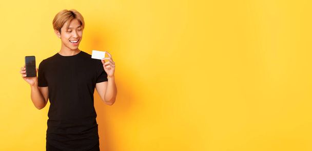 Guapo satisfecho asiático chico sosteniendo tarjeta de crédito y mostrando smartphone pantalla, sonriendo complacido, de pie amarillo fondo. - Foto, Imagen