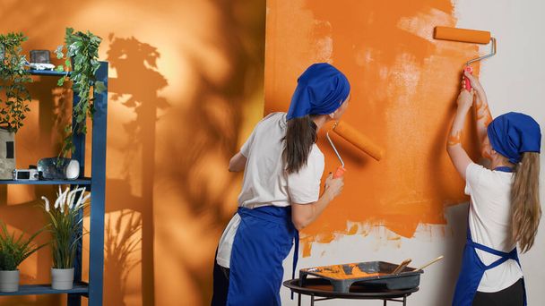 Οι άνθρωποι ζωγραφική τοίχους με πορτοκαλί χρώμα και βούρτσα κυλίνδρων, χρησιμοποιώντας εργαλεία και εξοπλισμό για να ζωγραφίσει το σπίτι εσωτερική διακόσμηση. Μητέρα και μικρό παιδί διασκεδάζουν αναδιακοσμώντας το διαμέρισμα. Σφηνάκι τρίποδα. - Φωτογραφία, εικόνα