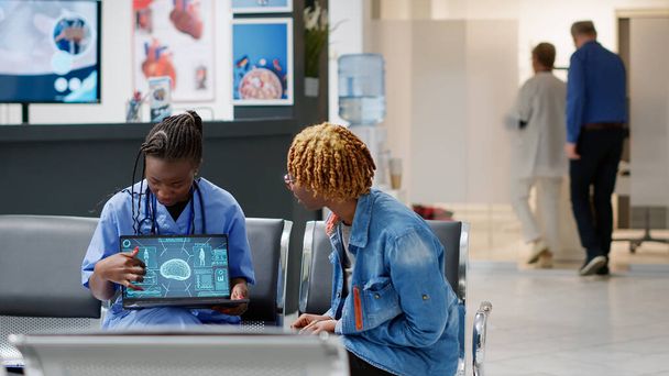 African american patient analyzing brain neurology scan at medical consultation in reception lobby with nurse. Woman and assistant looking at tomography and neural system diagnosis on laptop. - Foto, Bild