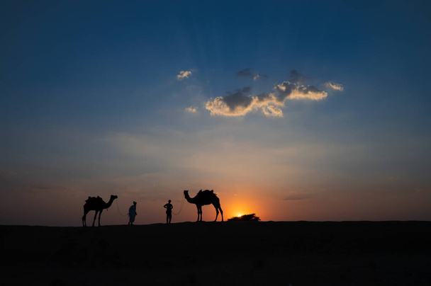 Silhouette de deux chameaux et de leurs chameaux dans les dunes de sable du désert de Thar, Rajasthan, Inde. Nuage avec coucher de soleil, ciel en arrière-plan. Cameleers font une vie hors de l'équitation de chameau par les touristes. - Photo, image