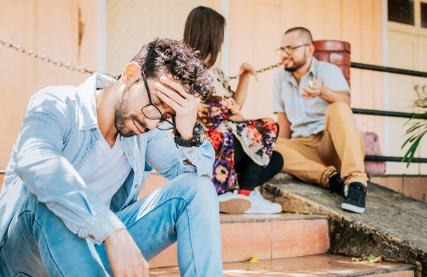 Sad man seeing his girlfriend cheating with his friend. Sad boyfriend sitting on some stairs while his girlfriend talking to another man. Girl talking to another man and her boyfriend is offended - 写真・画像