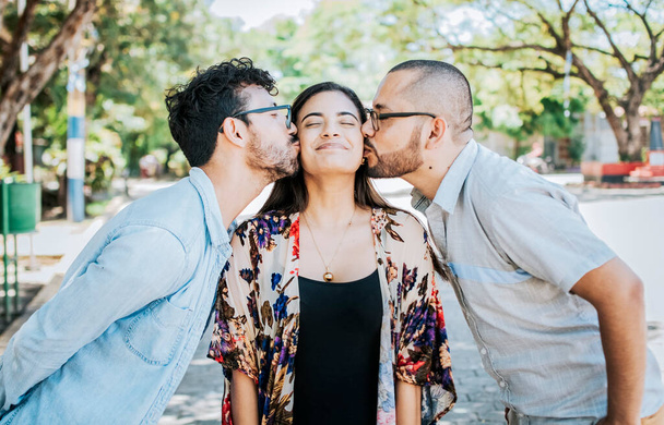 Love triangle concept. Polygamy concept. Two men kissing a girl cheek. Portrait of two guys kissing a girl cheek. Two young men kissing a woman cheek outdoor - Photo, Image