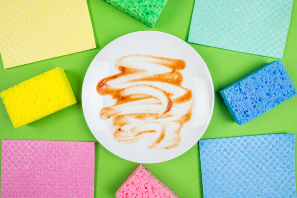 Photo from above of a dirty plate and multicolored cleaning pads on an isolated green background - 写真・画像