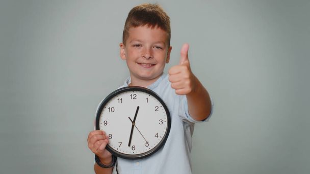 It is your time. Toddler kid boy showing time on wall office clock, ok, thumb up, approve, pointing finger at camera. Young teenager child schoolboy isolated alone on gray studio background indoors - Fotografie, Obrázek