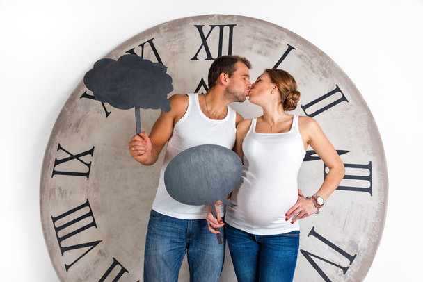 Happy Pregnant Couple dressed in white kissing and showing sign speech bubble banner looking happy excited and having idea on white background with giant clock. - Photo, Image