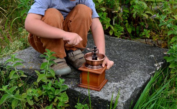 a little boy borrowed a manual coffee grinder and grinded biscuits. At my grandmother's cottage, she grinds with an old, historic wooden coffee maker with a brass handle. chubby boy in pants  kneeling - Foto, Imagen