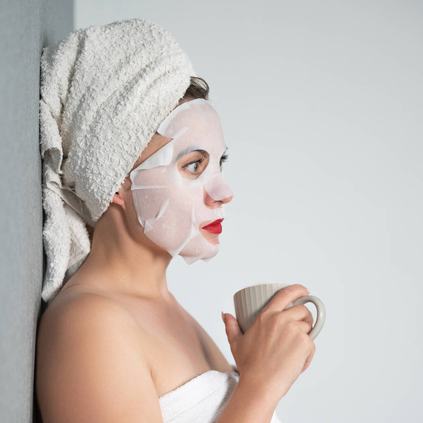 Facial care, a girl and a fabric mask on her face, a woman holds a cup with a drink in her hands. - Photo, Image