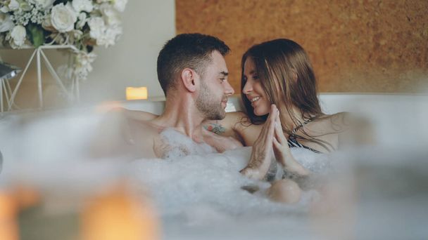 Newly married attractive couple is relaxing in jacuzzi kissing, touching hands, talking and laughing. Romantic relationship, passionate honeymoon and wellness concept. - Photo, image