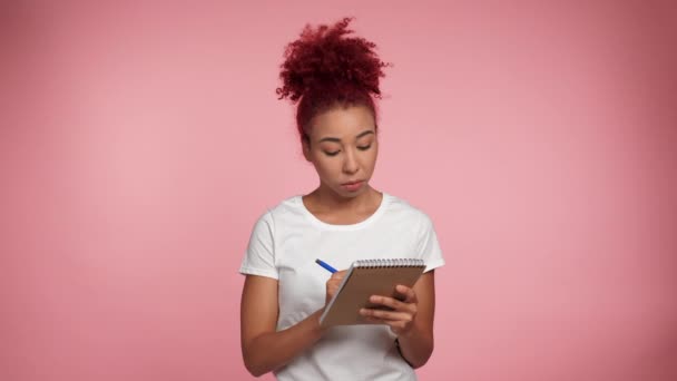 Gondolkodó mosolygós kreatív afro-amerikai nő vörös hajú jegyzetel papír notebook, írás kreatív ötlet, jövőbeli tervek és tennivalók listája, látszó ihlette elszigetelt rózsaszín háttér - Felvétel, videó