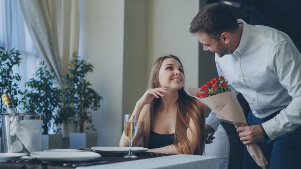 Happy girlfriend is getting flowers and present from her loving boyfriend after waiting for him alone in restaurant. Romantic relationship, gifts and fine dining concept. - Foto, afbeelding