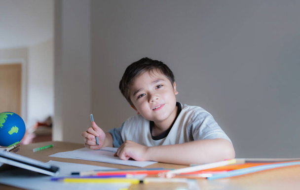 Хлопчик сидить на столі, малює і малює на білому папері, початковій школі і домашній школі, концепції дистанційної освіти.  - Фото, зображення