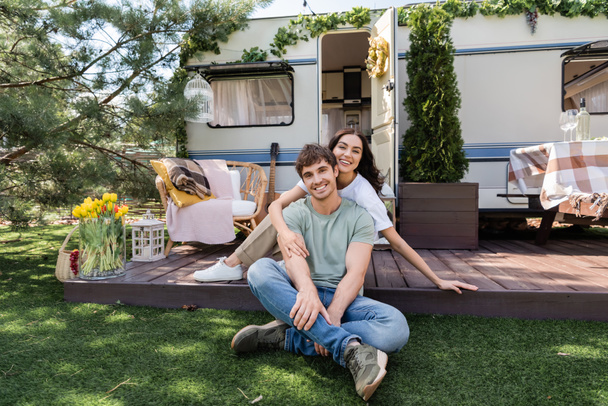 Smiling woman hugging boyfriend and looking at camera on terrace of camper van  - Foto, afbeelding