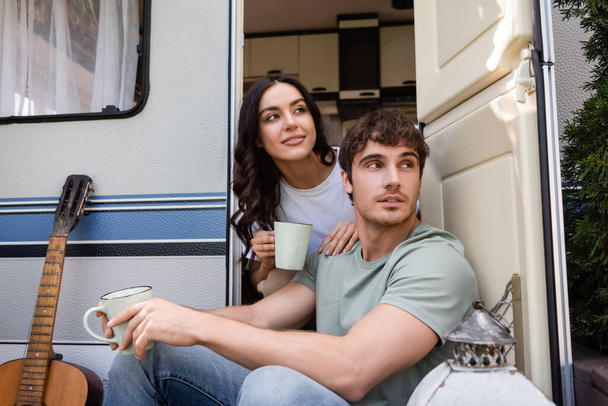 Young couple holding cups of coffee near acoustic guitar and camper van outdoors  - Foto, Imagem