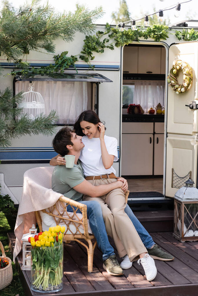 Smiling woman holding cup while sitting on boyfriend near camper van  - Foto, Imagen