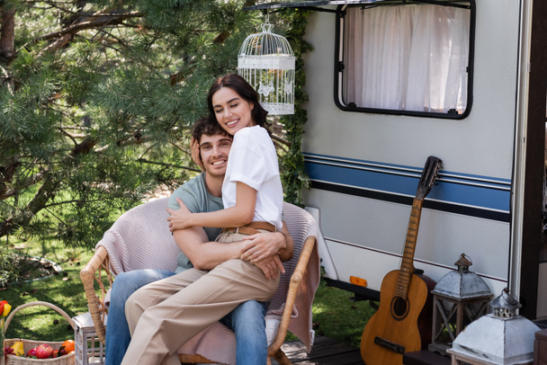 Positive woman hugging young boyfriend while sitting on armchair near camper van  - Photo, image