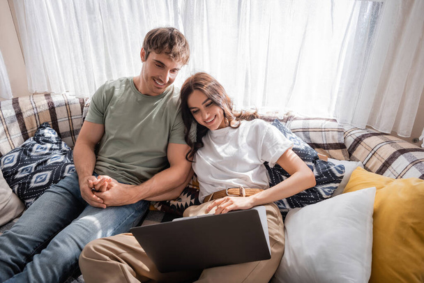 Smiling man holding hand of girlfriend using laptop on bed in camper  - Фото, изображение