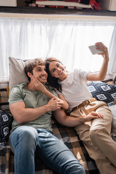 Smiling woman taking selfie with boyfriend on bed in camper  - 写真・画像