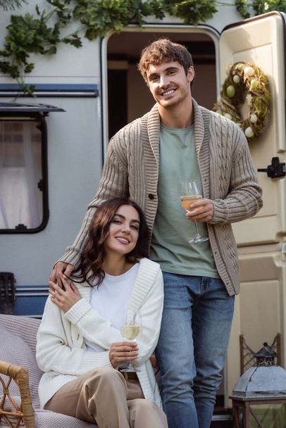 Young couple in warm cardigans holding wine and smiling at camera near camper van  - Valokuva, kuva