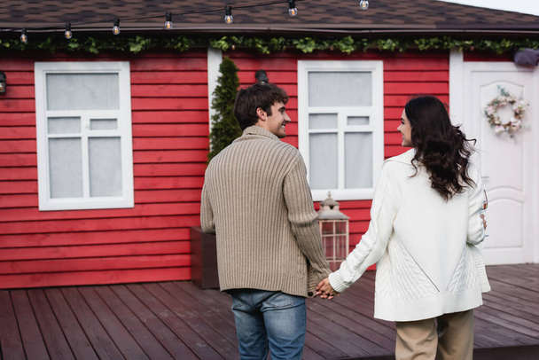 Side view of smiling couple in cardigans holding hands near blurred house  - Foto, Imagem