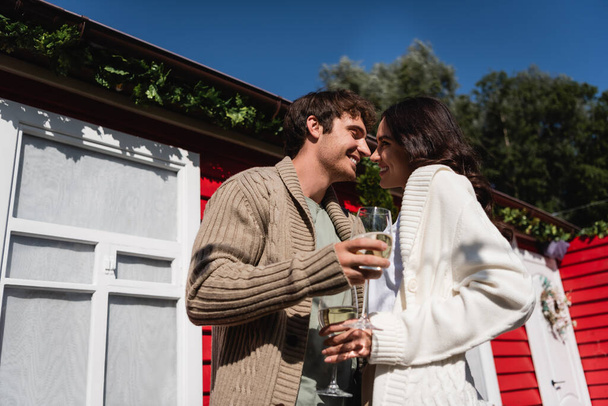 Low angle view of smiling young couple in cardigans holding glasses of wine near house outdoors  - Photo, Image