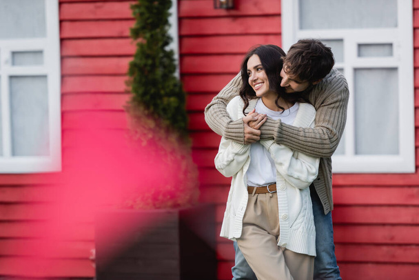 Man in warm cardigan hugging smiling girlfriend near house  - Foto, Imagem